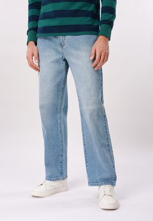 Mann iført lyseblå jeans med rette ben, hvite joggesko og en langermet skjorte med mørkegrønne og marineblå striper, stående foran en ensfarget bakgrunn.