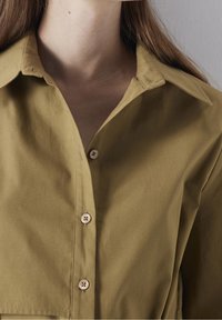 Primer plano de una persona que lleva una camisa de botones verde oliva con cuello, mostrando el cuello y la parte superior del pecho.