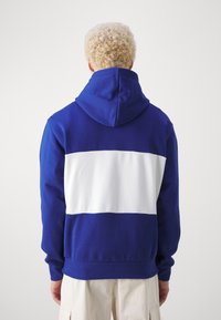 Hoodie w niebieskim kolorze z białym poziomym paskiem na plecach. Posiada teksturowany materiał i ściągacze przy mankietach.