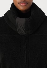 Schwarz gefütterte Jacke mit hohem Kragen. Weiche Textur, mit einem glatten schwarzen Stoffakzent am Rand des Kragens und einem gestickten Design.