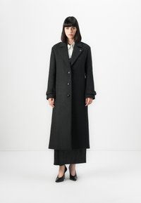 Woman standing against white background wearing long dark gray buttoned coat, black skirt, white shirt, and black heels.