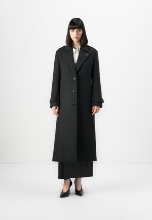 Donna in piedi contro uno sfondo bianco, che indossa un lungo cappotto grigio scuro con bottoni, una gonna nera, una camicia bianca e tacchi neri.