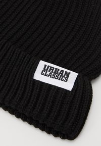 Čierna pletená čiapka s rebrovanou textúrou. Biela nálepka na prednej strane zobrazuje "URBAN CLASSICS" tučným čiernym písmom.