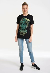 Black T-shirt featuring a Slytherin crest design in green, accented by serpents. Soft cotton material with a relaxed fit. Paired with light blue jeans.