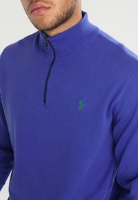Gilet zippé violet en tissu tricoté. Il dispose d'un col montant et d'un petit logo vert sur la poitrine gauche.