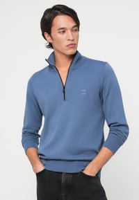 Blauwe gebreide pullover met een kwart-rits kraag, geribde manchetten en onderrand, met een klein logo op de borst. Gemaakt met een donker paar broek erbij.