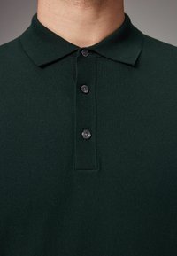 Polo de color verde oscuro hecho de tejido de punto, con un cuello tipo polo y tres botones negros en la tapeta. Textura suave.