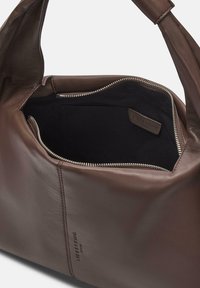 Brązowa skórzana torebka o gładkiej fakturze, z zamkiem błyskawicznym na górze i czarnym materiałowym wnętrzem. Posiada małe logo z boku.