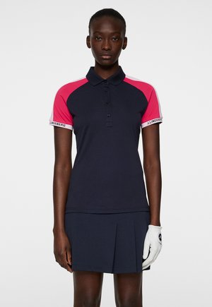 Dunkelblauses Poloshirt mit rosa Raglanärmeln, Drei-Knopf-Leiste und gebrandeten Akzenten; kombiniert mit einem dunkelblauen Faltenrock und einem weißen Golfhandschuh.