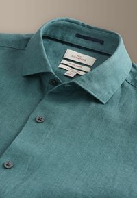 Chemise en lin vert sarcelle à boutons, col pointu, boutons noirs, et étiquettes intérieures indiquant la taille M, 100 % lin et coupe régulière.