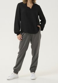 Blusa negra de manga larga con escote en V y puños fruncidos, combinada con pantalones grises ajustados con cintura elástica y zapatillas blancas.