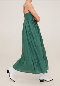 Zelené maxi šaty s nastavitelnými ramínky, nabíraným lemem a splývavým vzhledem. Nosené s bílými kotníkovými botami na podpatku.