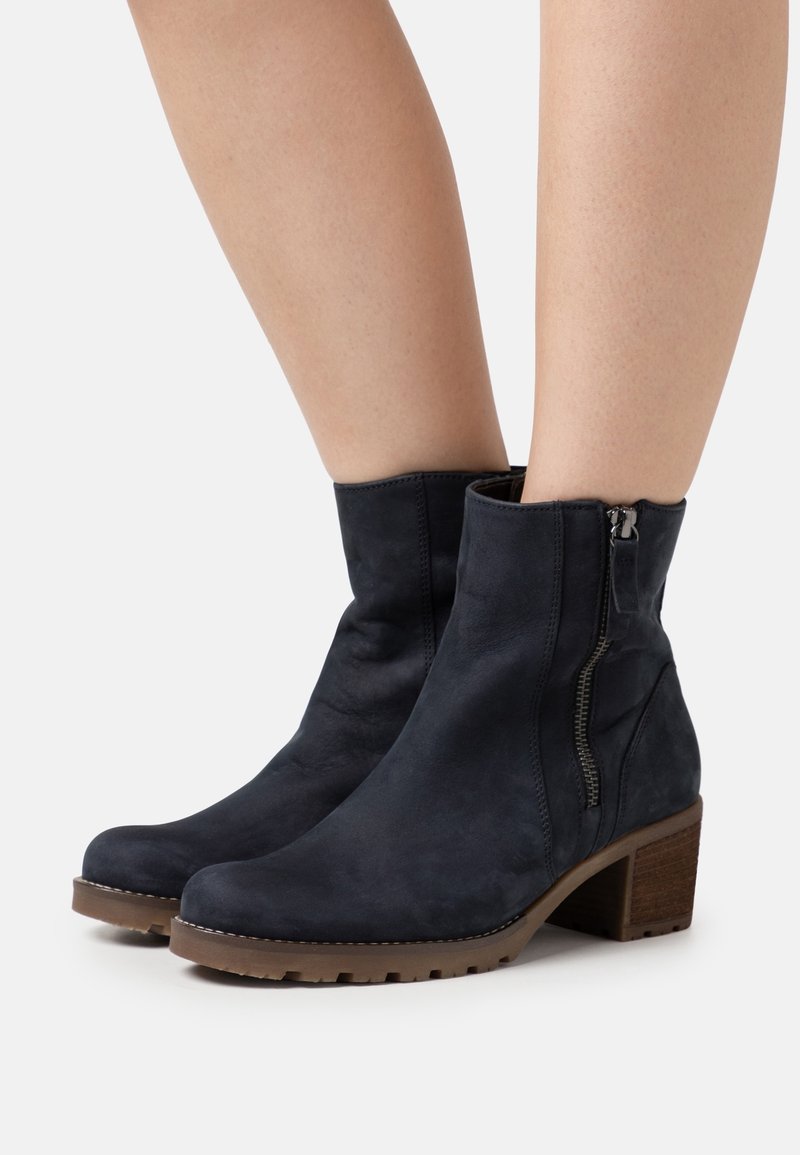 Bottines en daim noir à fermeture éclair latérale, talon bloc et semelle en caoutchouc marron portées sur jambes nues.