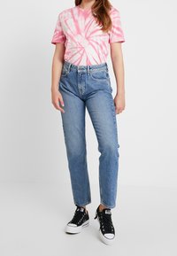 Camiseta de tie-dye en tonos rosa y blanco combinada con jeans azul de cintura alta y zapatillas de lona negras. Look casual con un ajuste relajado y puños enrollados.