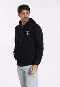 Haut de survêtement noire zippée avec une poche kangourou, capuche à cordon et patch logo. Associé à un pantalon de survêtement gris clair. Texture de tissu lisse.