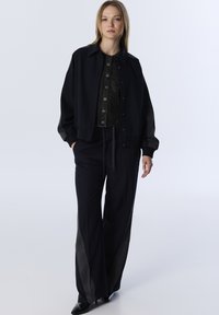 Zwarte bomberjack met drukknoopjes, gecombineerd met een donker overhemd met knopen en wijde broek met een grijze zijsstrijpe. Soepele textuur.