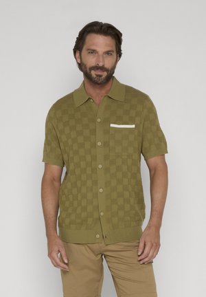 Mand iført olivengrøn kortærmet skjorte med ternet mønster og beige bukser, stående mod en ensfarvet lys baggrund.