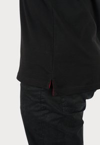 Tricou negru cu fante laterale, având detalii de cusătură roșie la tiv. Purtați cu pantaloni texturați negri.