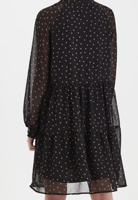 Černé šaty s puntíky, dlouhými průhlednými rukávy, vícestupňovým designem a kulatým výstřihem. Materiál se zdá být lehký s splývavou texturou.