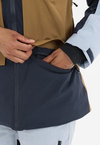 Vedenkestävä takki kullankeltaisena ja laivastonsinisenä, jossa on vetoketjullinen tasku. Ominaisuuksia ovat sileä pinta ja kerroksellinen muotoilu, jossa on kontrastoivat värit.