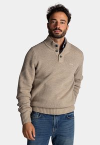 Beige gebreide trui met een hoge kraag, ribbelmanchetten en onderrand. Bevat twee donkere knopen en een gestructureerd oppervlak. G gedragen over een geruite shirt.