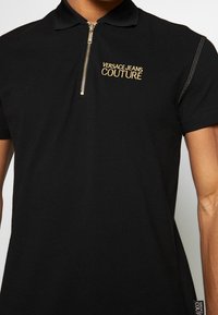 Zwarte polo shirt van katoen met een ritskraag. Voorzien van gouden borduring "VERSACE JEANS COUTURE" en zijnaad details.