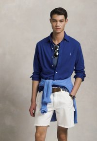 Model draagt een blauwe langarmige shirt, gestreepte t-shirt eronder, witte shorts en een lichtblauwe sweater om de taille gebonden.