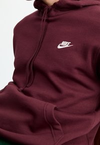 Vínově červená mikina s kapucí z měkkého materiálu, s kapucí na stažení a malým bílým logem Nike na hrudi. Součástí je kapsa typu klokánek.
