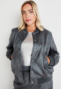 Grå bomberjakke i myk, teksturert stoff med trykknapp foran, ribbede mansjetter og sidelommer, båret over en sølvtop.