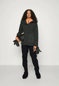 Γυναίκα με σγουρά μαλλιά, φορώντας μαύρο παντελόνι και σακάκι πάνω από πορτοκαλί τοπ, κρατώντας μια μαύρη κράνος και γάντια, στέκεται μπροστά σε απλό φόντο.