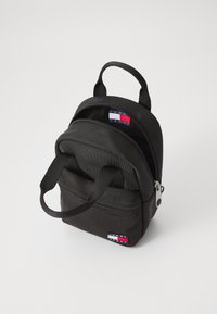 Mini sac à dos noir en tissu durable, comprenant deux compartiments zippés, des poignées courtes et une étiquette de logo aux couleurs rouge, blanc et bleu.