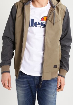 Homme portant une veste à capuche beige et grise sur un t-shirt blanc graphique et un jean bleu, debout les mains détendues le long du corps.