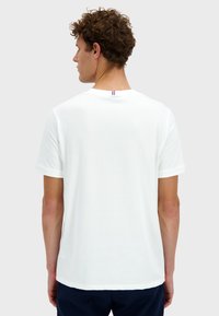 Bílá bavlněná tričko s krátkými rukávy, kulatým výstřihem a malým červeným, bílým a modrým pruhovaným akcentem na zadní části.