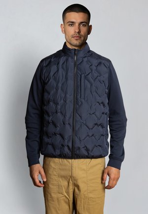 Man wearing navy blue quilted zip-up vest over long-sleeve shirt with tan pants, standing against plain light gray background.