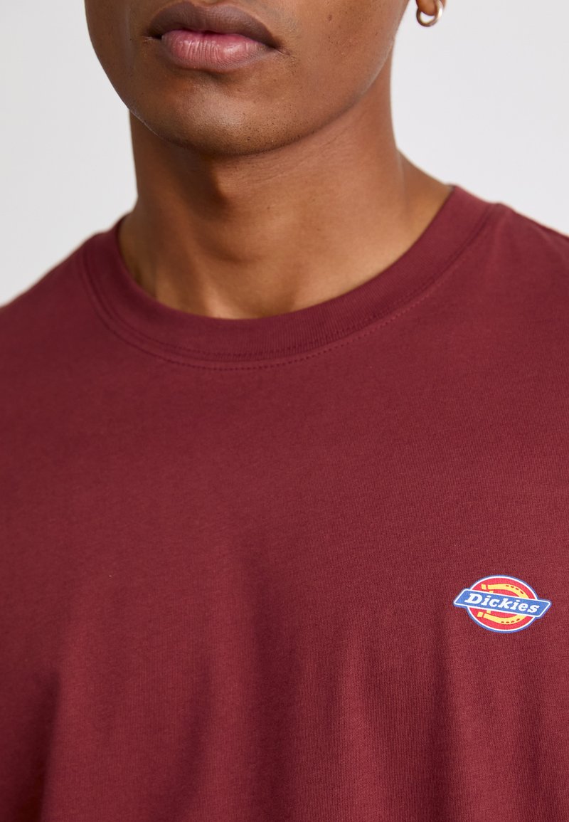 Bordeaux katoen T-shirt met een ronde hals en een klein rond "Dickies" logo in blauw, rood en geel op de borst. Soepele textuur.