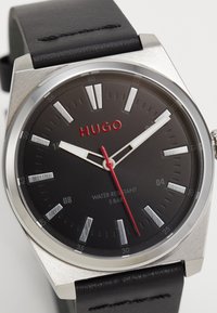 Reloj de cuero negro con caja de acero inoxidable plateado, esfera negra, acentos rojos, marcadores de hora plateados y resistencia al agua con una clasificación de 5 bares.
