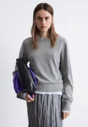 Gray sweater with a crew neckline, worn over a white shirt. Holding a black bag and purple garment, textured gray skirt visible.
