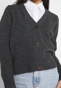 Grauer gestrickter Cardigan mit V-Ausschnitt, drei großen braunen Knöpfen und gerippter Textur, getragen über einem weißen Hemd mit Kragen.