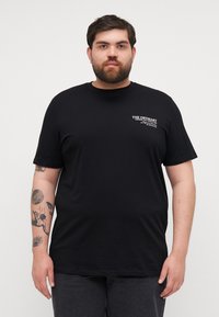 Black cotton t-shirt with short sleeves, round neckline, and a white logo on the left chest. Features a relaxed fit and soft texture.