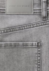 Gros plan sur un jean en denim gris clair montrant une poche arrière, une passoire de ceinture et une étiquette rectangulaire cousue portant l'inscription "STREET ONE STUDIO".
