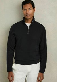 Polo de media cremallera negro en tela suave con puños y bajo acanalados. Cuenta con una cremallera plateada y un forro interior blanco, combinado con pantalones claros.