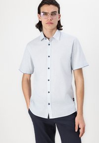 Chemise à manches courtes bleu clair avec un fini texturé, boutons foncés, biais contrastant bleu marine et une petite étiquette logo sur l'ourlet.