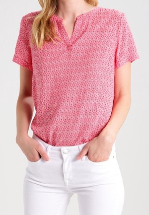 Frau trägt eine kurzärmelige, rosa gemusterte Bluse, die in hochgeschnittene weiße Hosen gesteckt ist, Hände in den Taschen ruhend.