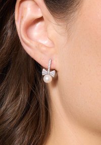 Srebrne kolczyki z akcentem w kształcie kokardy, ozdobione cyrkoniami oraz centralną okrągłą perłą, umieszczone na tle teksturowanej skóry.