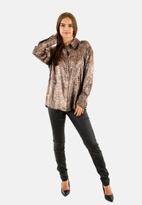 Camisa de bronce con patrón de piel de serpiente, con cuello, mangas largas y cierre de botones, combinada con pantalones negros ajustados y tacones negros afilados.