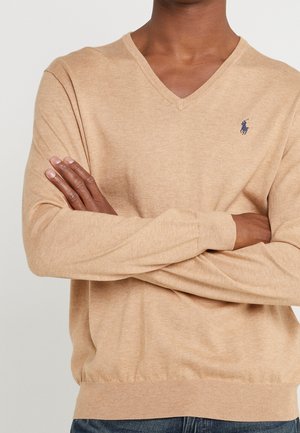 Mężczyzna z założonymi rękami, ubrany w beżowy sweter z dekoltem w serek i małe granatowe logo gracza polo na lewej piersi.