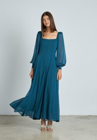 Teele langes Kleid mit puffierten, transparenten Ärmeln, quadratischem Ausschnitt und fließendem Stoff. Getragen mit verzierten, offenen High Heels auf einer hellen Oberfläche.