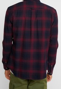 Persona che indossa una camicia a maniche lunghe rossa e blu a quadri, vista da dietro, abbinata a pantaloni cargo verde oliva.