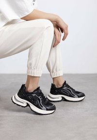 Zapatillas deportivas negras con parte superior sintética texturizada, media suela blanca y suela de goma, combinadas con pantalones jogger de color beige claro.