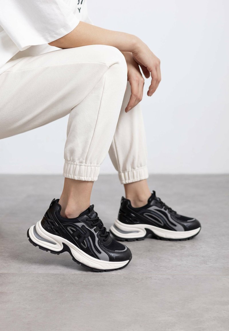 Zapatillas deportivas negras con parte superior sintética texturizada, media suela blanca y suela de goma, combinadas con pantalones jogger de color beige claro.